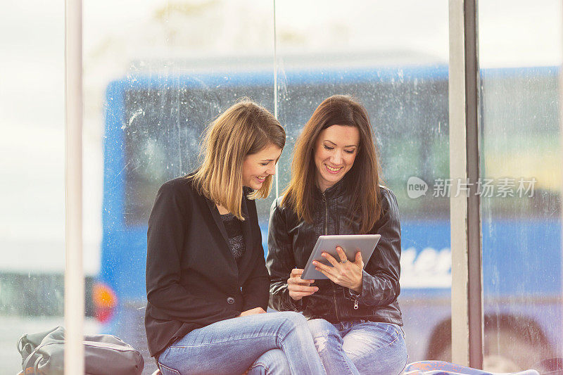 Two friends using digital tablet on bus station